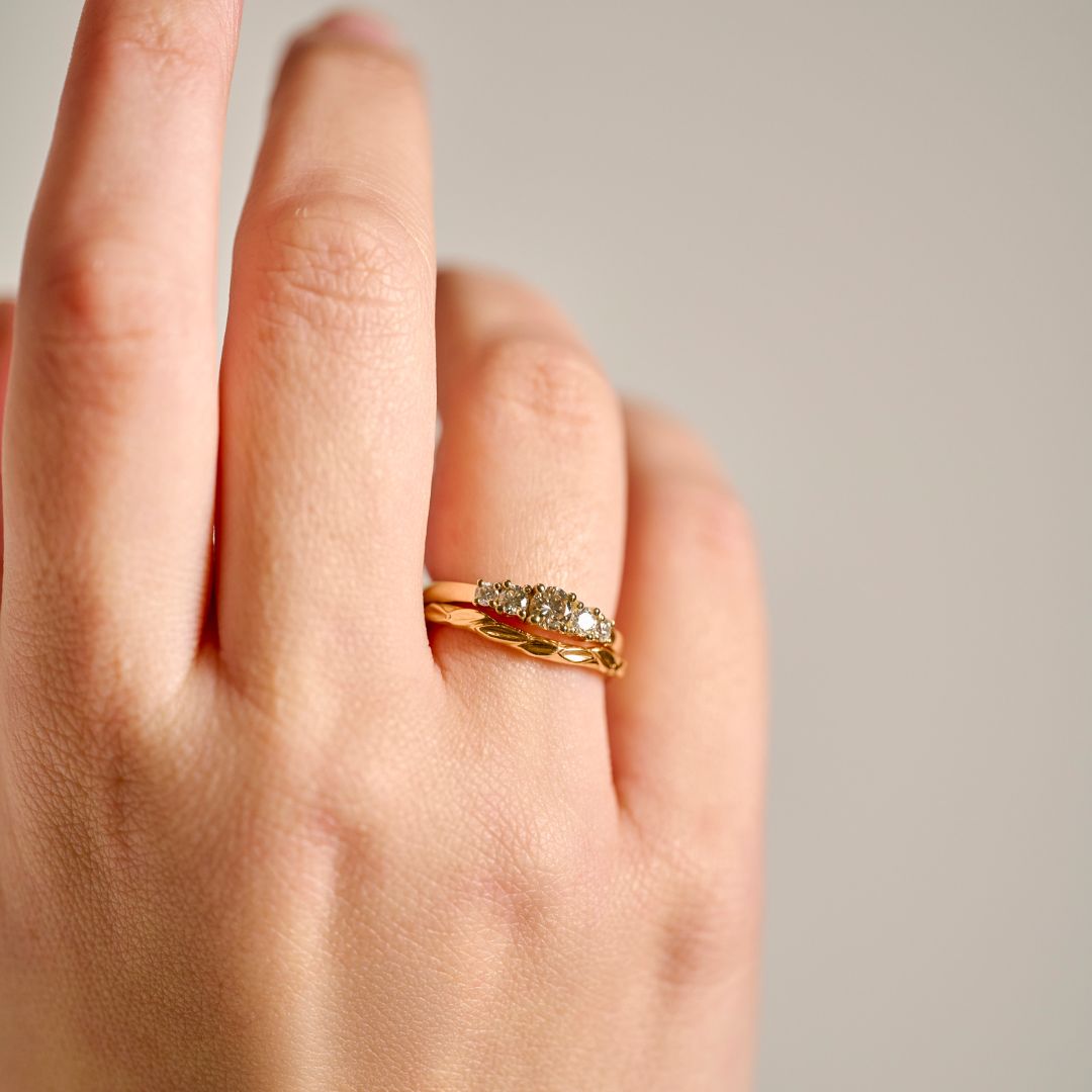 A close-up photograph of a woman's hand wearing an organic leaf wedding ring stacked with a cognac diamond engagement ring, set against a soft  grey out of focus background.