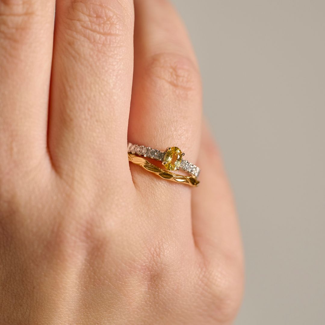 A close-up photograph of a woman's hand wearing an organic leaf wedding ring stacked with a yellow sapphire and diamond engagement ring , set against a soft neutral background.