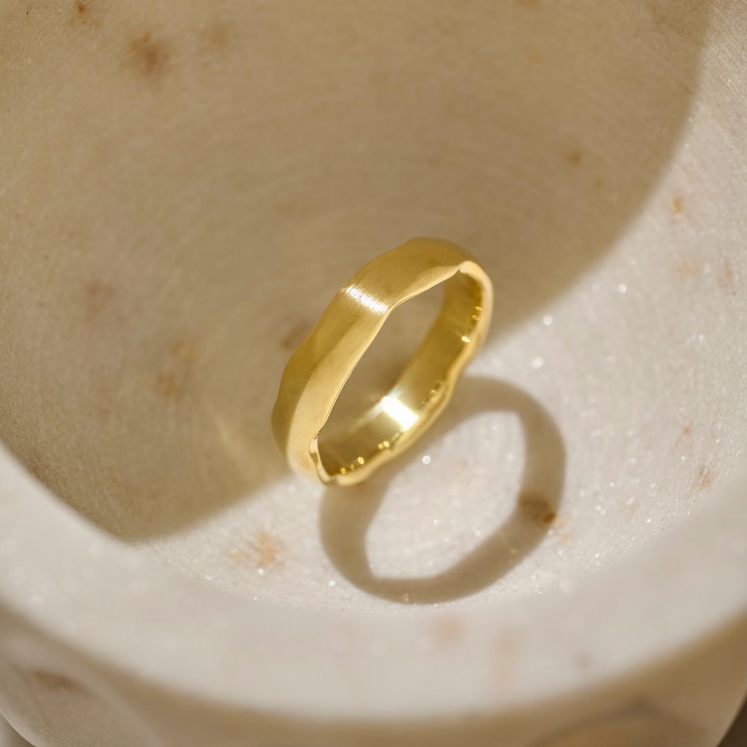 A minimalist matte yellow gold mens organic wedding ring resting inside a textured, light beige natural stone bowl, casting a soft shadow.