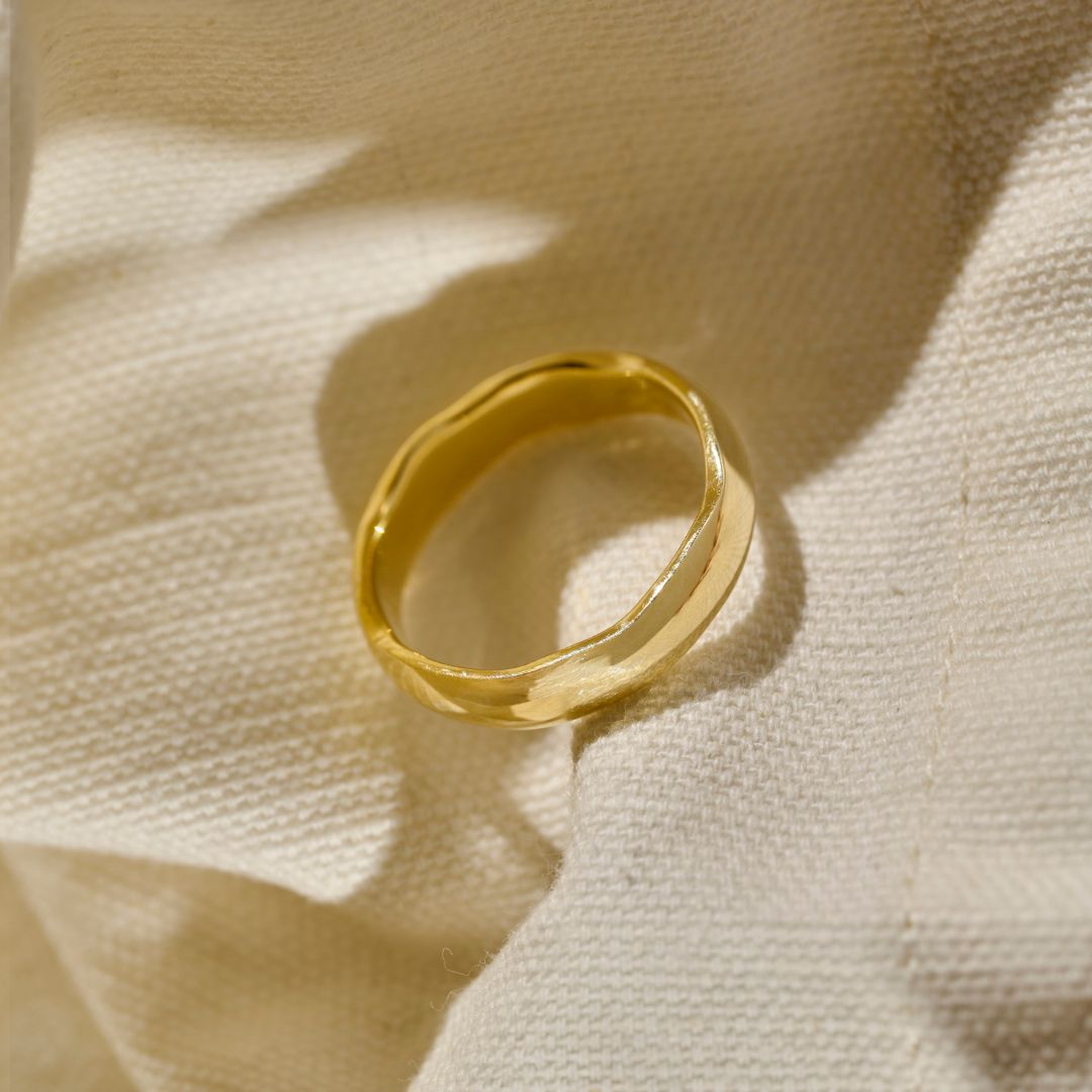 A polished yellow gold mens wedding  ring with a slightly organic, wavy shape, resting on a textured cream-coloured fabric cloth.