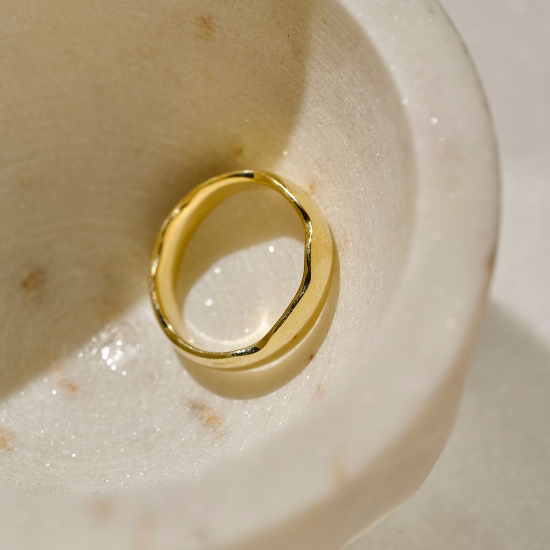 A polished yellow gold statement ring with an organic, wavy band shape photographed inside a textured cream-coloured stone dish.