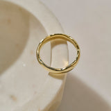 A polished yellow gold ring with a slightly wavy, organic silhouette, positioned on its side and resting on the edge of a cream-coloured stone bowl.