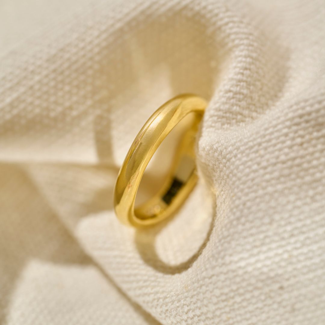 A polished yellow gold wedding ring with a slightly wavy, organic band shape, photographed on a textured cream woven fabric napkin with soft shadows.