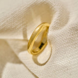 A polished yellow gold wedding ring with a slightly wavy, organic band shape, photographed on a textured cream woven fabric napkin with soft shadows.