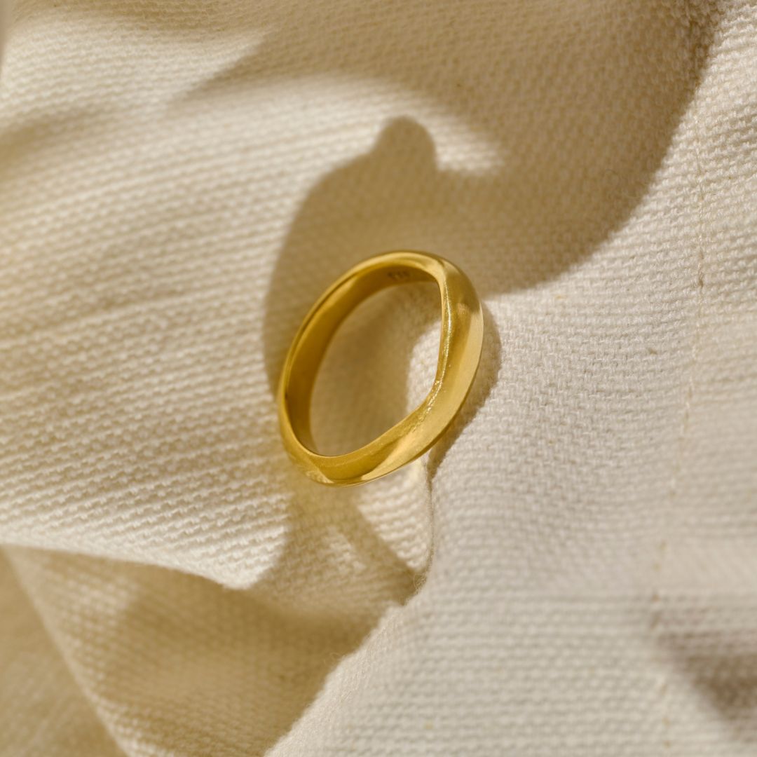 A polished yellow gold wedding ring with a slightly wavy, organic band shape, leaning on a textured beige woven fabric cloth with soft shadows.
