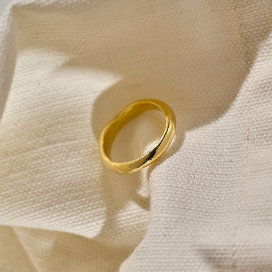 A polished yellow gold wedding ring resting on its side with a slightly wavy, organic band shape, positioned on a beige woven fabric material with soft shadows.