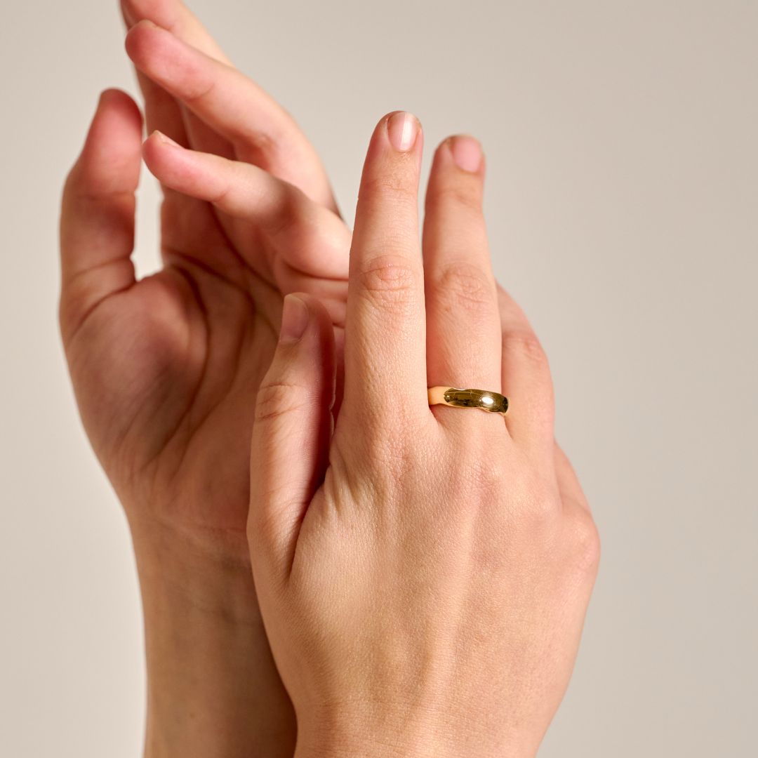 A close-up of woman's hands against a warm beige background, with the right hand wearing a statement organic yellow gold ring on her middle finger.