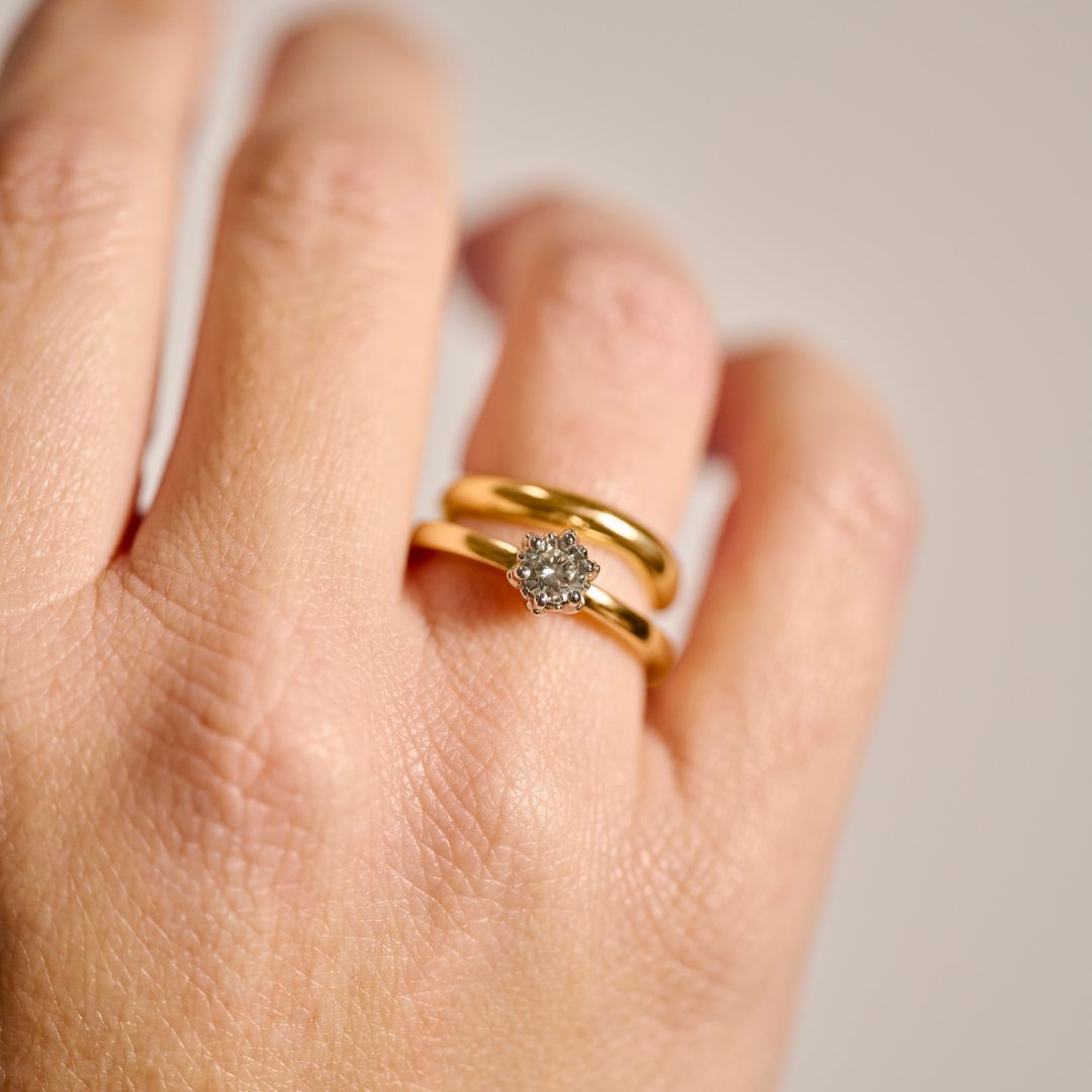 A close-up of a woman's hand wearing a gold ring featuring a round natural diamond in a Protea floral setting stacked with an organic yellow gold organic wedding ring, photographed against a soft light grey background.