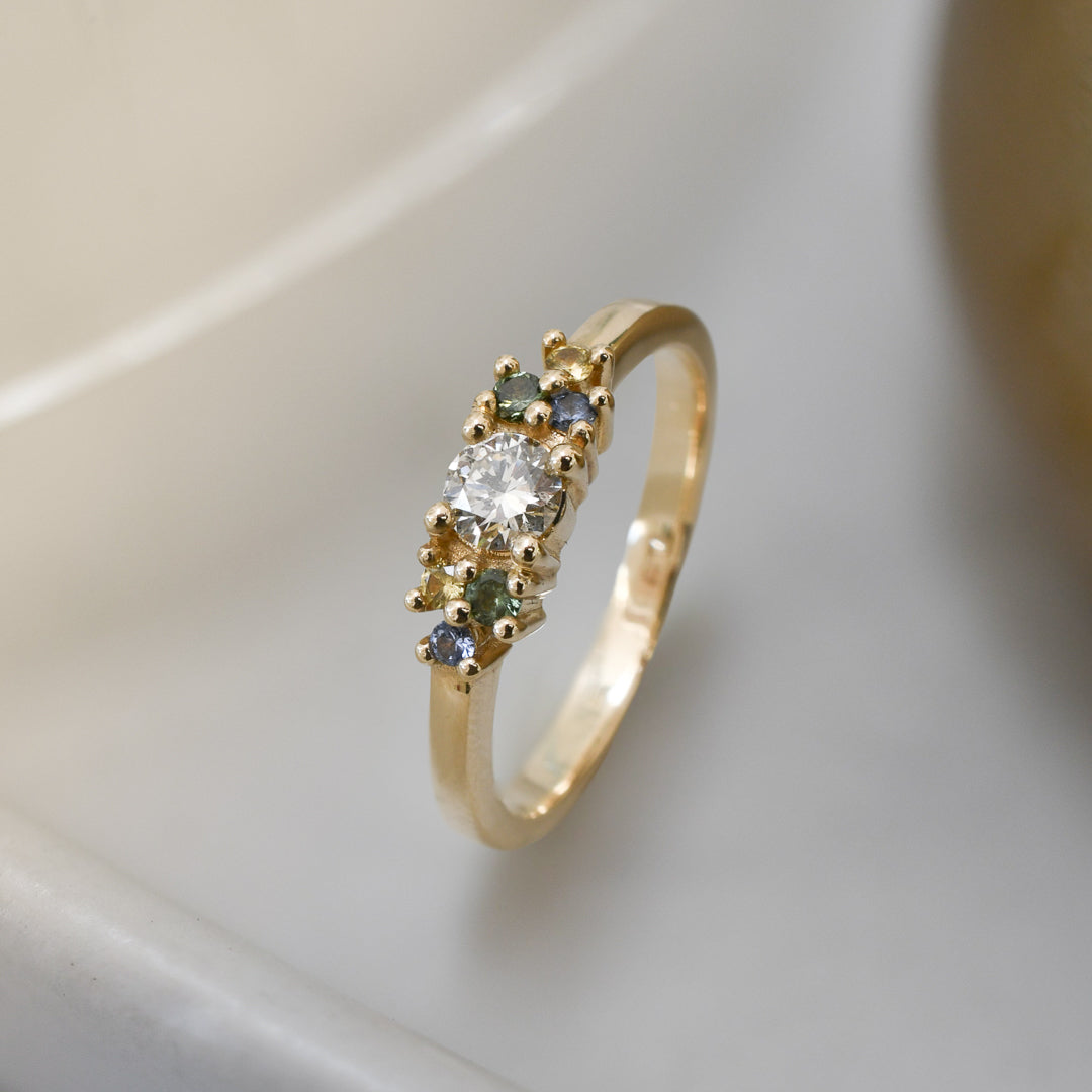 Yellow gold cluster ring set with a central natural diamond and additional blue, green, and yellow sapphires on either side. on a neutral background