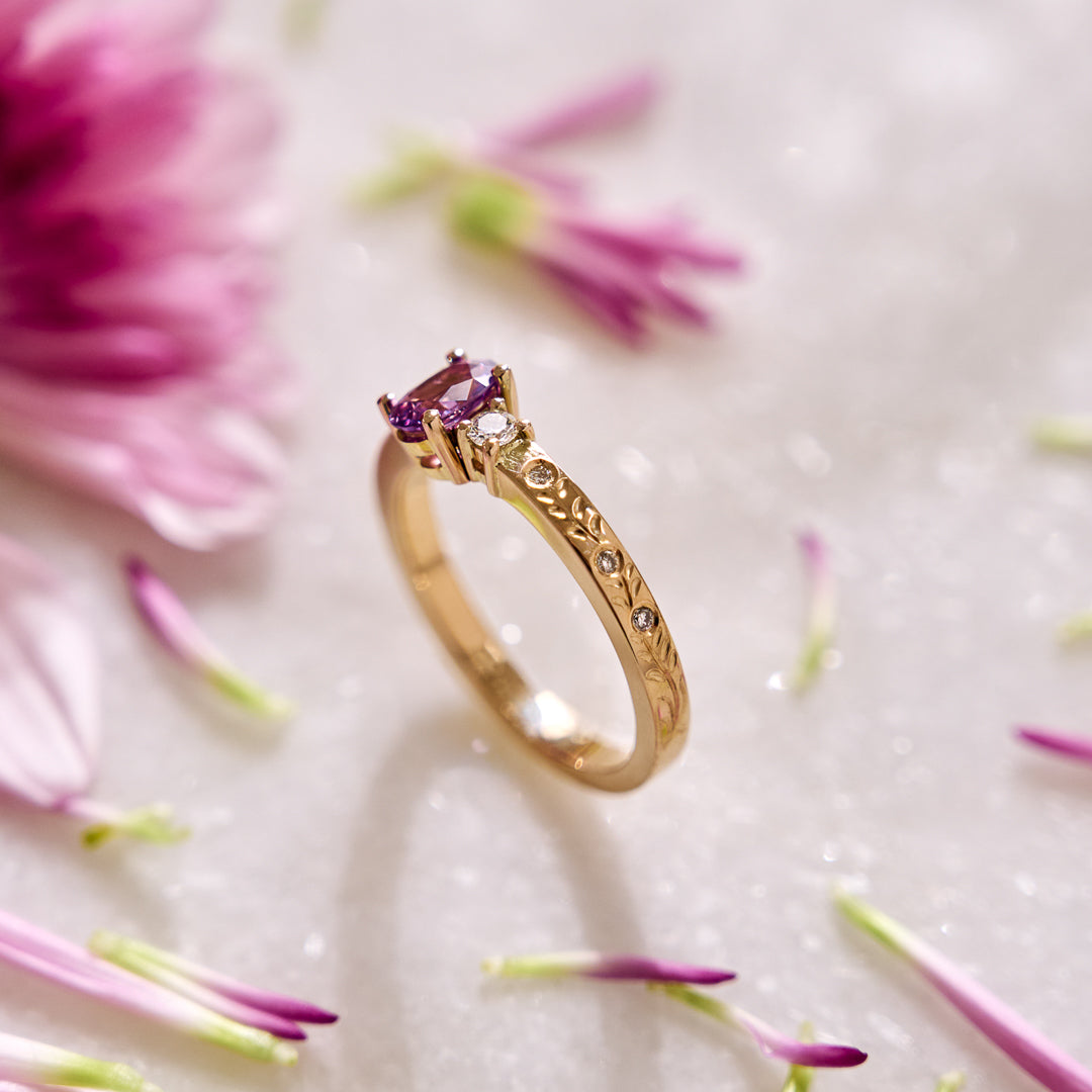 Yellow gold ring set with a purple sapphire and natural diamonds, featuring lavender-inspired hand engraving on the shank with natural diamonds on a white background with pink flower petals