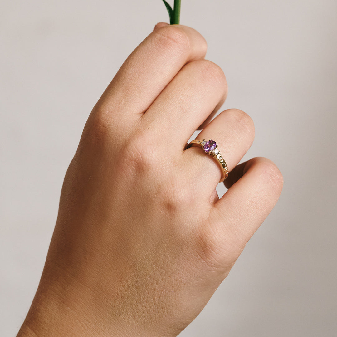 Hand wearing a ring with a purple sapphire and natural diamonds holding a small plant on a neutral background