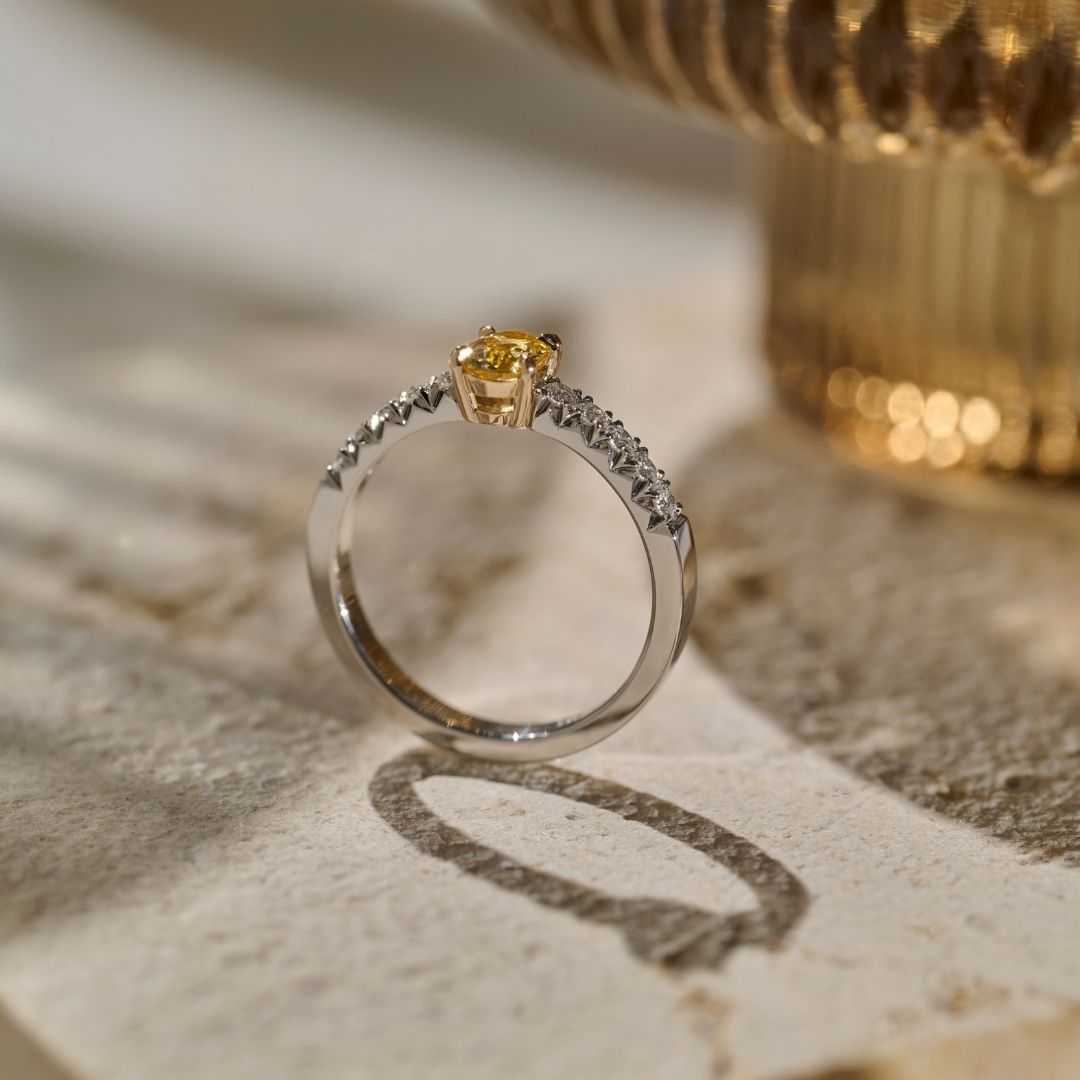 Side view photograph of a oval yellow sapphire set in a four claw yellow gold setting and a platinum shank with pavé set round colour diamonds, resting on a textured beige stone surface.